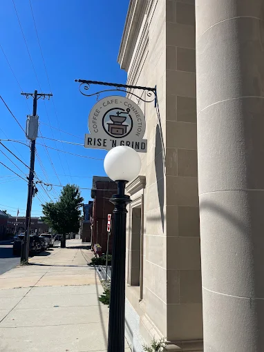 The Rise 'N Grind sign hanging from the stone facade of the old bank building on North Street, next to a round streetlamp